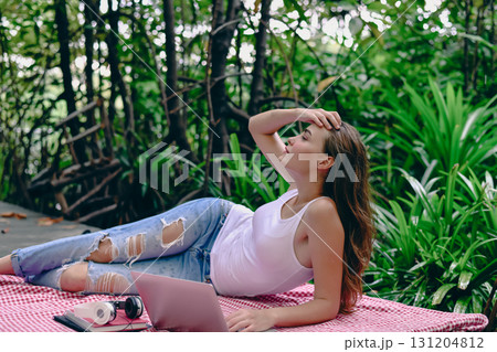 A young woman relaxes outdoors, lying on a blanket, enjoying nature while using a laptop amidst lush greenery. A young woman relaxes outdoors, lying on a blanket, enjoying nature while using a laptop amidst lush greenery. 131204812