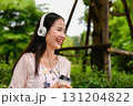 A young woman enjoys her coffee while wearing headphones outdoors, radiating joy in a serene green environment. 131204822