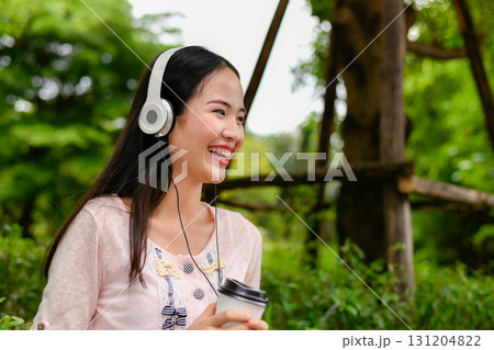 A young woman enjoys her coffee while wearing headphones outdoors, radiating joy in a serene green environment. 131204822