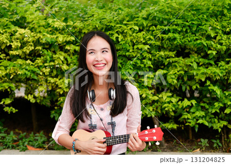 Joyful girl playing ukulele outdoors, radiating happiness in a lush green environment. 131204825