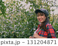 Cheerful young woman enjoying nature, wearing a cap and plaid shirt, smiling amidst lush greenery. 131204871