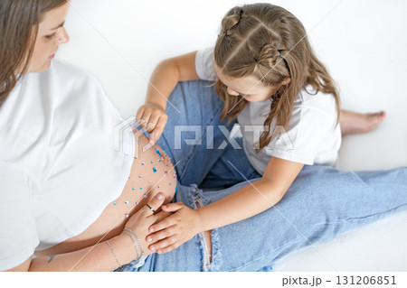 The daughter examines the rhinestones on the pregnant mother's stomach. A girl with her mother on a white studio background. Waiting for the baby to be born. 131206851
