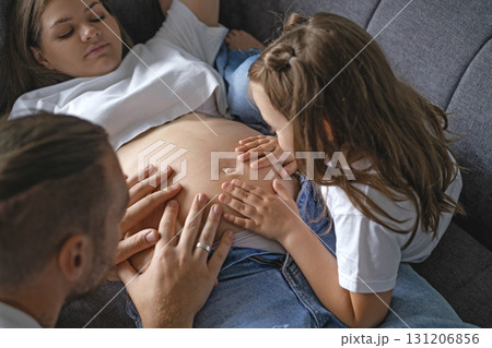 A mother pregnant with second child is lying on the couch with her youngest daughter and her husband, surrounded by love and care. The father touches the belly and feels the baby's thrusts in womb 131206856