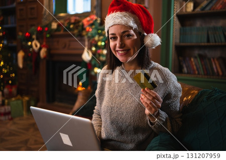 Merry Christmas. Young woman shopping online with laptop paying with gold credit card for Christmas gifts. Girl buying on Internet enter credit card details enjoying winter holidays at home 131207959