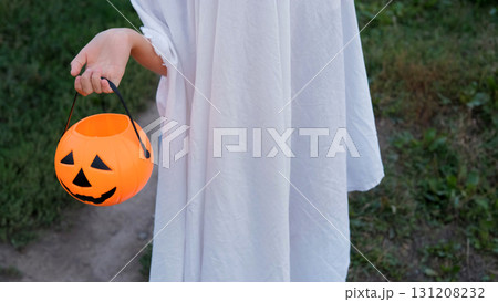 A young girl dressed in a ghost costume standing on an autumn path holding a pumpkin shaped basket filled with candy Halloween celebration concept 131208232