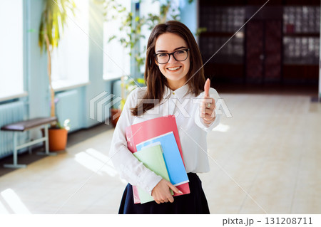 Happy student standing indoor in the university building looking to the camera showing thumbs up and holding books, exercise books in her hands 131208711