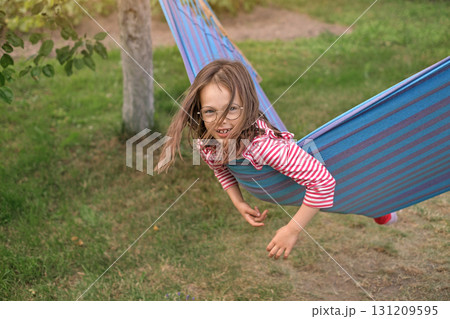 A funny girl with glasses is swinging on a blue striped hammock in the garden on a summer day. Summer vacation vacation 131209595