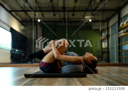 Young woman lying on yoga mat hugging knees to chest in relaxation pose 131211500