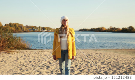 Happy young woman standing on sunny beach looking away, wear jacket, sea coast background Happy young woman standing on sunny beach looking away, wear jacket, sea coast background 131212488