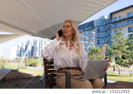 Woman engaged in a business call while working on her laptop in a modern outdoor setting during a 131212933