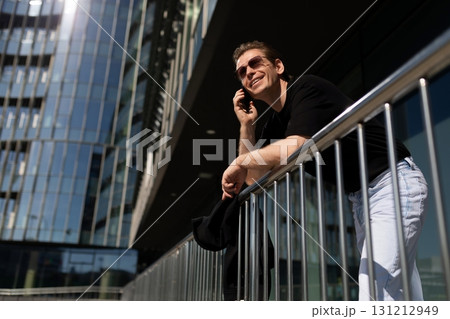 Man talking on the phone while leaning on a railing outside a modern building on a sunny day 131212949