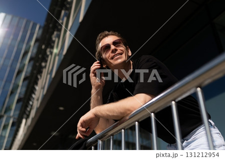 Man talking on a mobile phone while leaning on a railing outside modern buildings on a sunny day 131212954