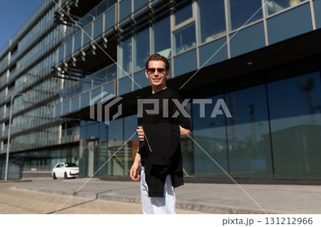 Young man enjoys a sunny day outside a modern building while casually holding a jacket 131212966
