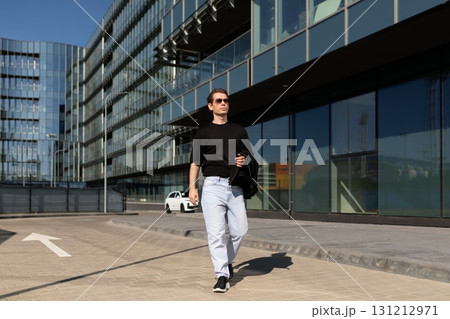 Urban man walks confidently in modern city area during sunny day while holding jacket Urban man walks confidently in modern city area during sunny day while holding jacket 131212971