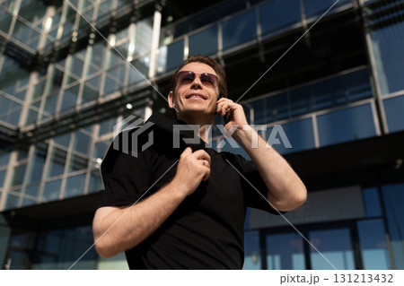 Man in sunglasses smiles while using phone outside modern building during sunny day 131213432