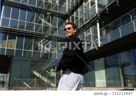 Young man smiles confidently while posing outside a modern glass building during a sunny day 131213447