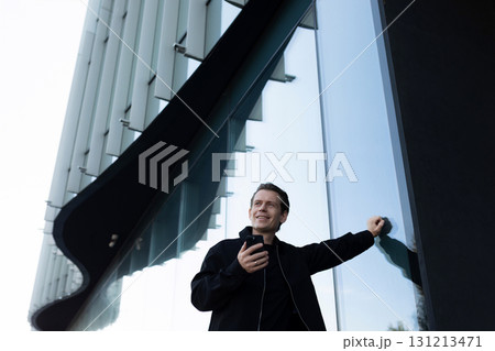 Smiling man standing near modern building while using smartphone in daylight 131213471