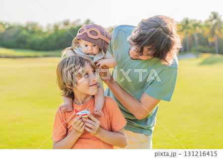 Father, son, and newborn on a meadow. Baby wearing a pilot hat, symbolizing dreams of aviation. Concept of family bonding, future pilot, and childhood aspirations Father, son, and newborn on a meadow. Baby wearing a pilot hat, symbolizing dreams of aviation. Concept of family bonding, future pilot, and childhood aspirations 131216415