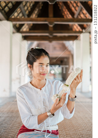 A woman in traditional Thai attire holds a jasmine garland, showcasing cultural heritage and elegance in a serene architectural setting. 131218822