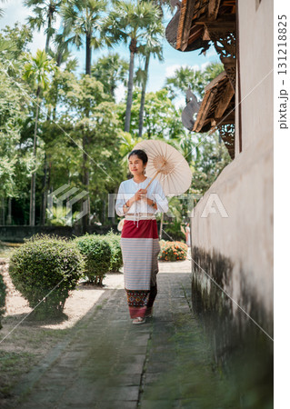 A woman in traditional attire walks with a parasol through a vibrant garden, surrounded by lush greenery and historic architecture. A woman in traditional attire walks with a parasol through a vibrant garden, surrounded by lush greenery and historic architecture. 131218825
