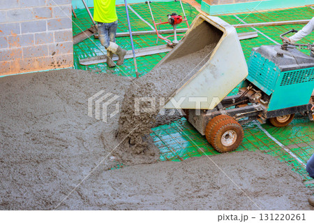 Workers are busy pouring concrete onto floor at construction site, uses wheeled concrete wheelbarrow during build. 131220261