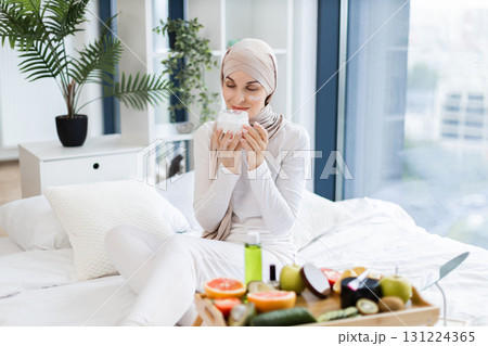 Young Muslim woman using moisturizer cream for self-care surrounded with fresh healthy ingredients. Promotes natural beauty, relaxation, and skincare. Young Muslim woman using moisturizer cream for self-care surrounded with fresh healthy ingredients. Promotes natural beauty, relaxation, and skincare. 131224365