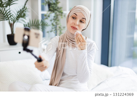 Young Muslim woman in light-colored hijab using cotton pads, blogging about skincare routine and personal health, sitting in an elegant and sunlit room while capturing content on a mobile device. Young Muslim woman in light-colored hijab using cotton pads, blogging about skincare routine and personal health, sitting in an elegant and sunlit room while capturing content on a mobile device. 131224369