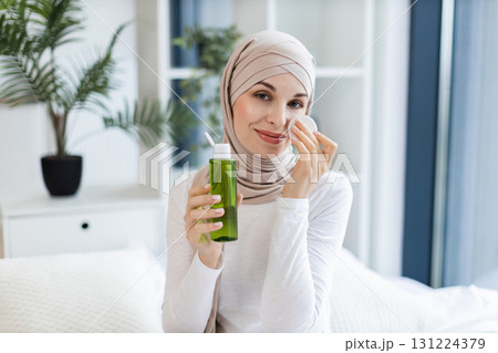 Young Muslim woman wearing beige hijab cleansing her face with lotion and cotton pad. Seated in modern bright home environment with plants in background, practicing skincare routine Young Muslim woman wearing beige hijab cleansing her face with lotion and cotton pad. Seated in modern bright home environment with plants in background, practicing skincare routine 131224379