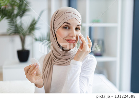 Young Muslim woman wearing hijab uses cotton pads to cleanse skin or remove makeup. Scene captures calm moment in airy home setting. Highlights skincare beauty routines lifestyle self-care and beauty. 131224399