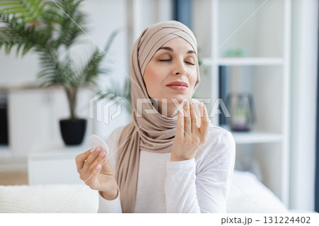 Young Muslim woman in hijab using cotton pads for skin cleaning, foreground showcasing vibrant and natural lifestyle concept. Scene reflects self-care, cosmetics, beauty, and peaceful indoor setting. 131224402