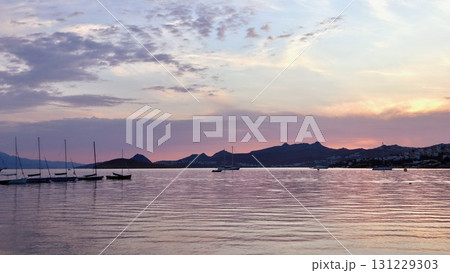 A peaceful coastal sunset with soft pink and purple clouds reflected on the calm water, with a silhouette of distant hills and a line of sailboats. 131229303