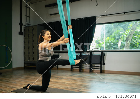 Smiling woman practicing aerial yoga using blue fabric hammock while stretching leg in fitness studio 131230971