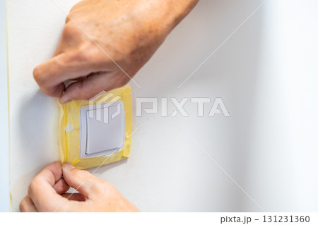 Man covering a light switch with masking tape before painting the wall white. Home renovation, surface protection and careful preparation during interior work. 131231360