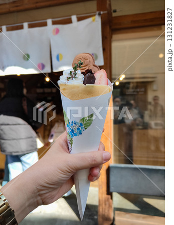 A hand holds a Japanese crepe filled with waffle cream, chocolate, and fruit, freshly served from a street food shop. The sweet dessert is wrapped in paper with a floral design, highlighting a casual 131231827