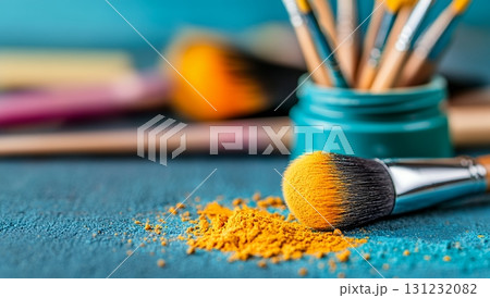 Golden makeup powder spills near a brush, showcasing vibrant hues against a teal background 131232082