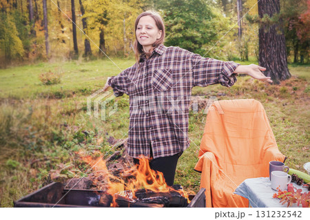 A smiling young woman in a brown plaid shirt enjoys an autumn picnic in nature around a campfire in autumn. A smiling young woman in a brown plaid shirt enjoys an autumn picnic in nature around a campfire in autumn. 131232542