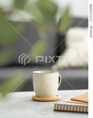 Ceramic coffee mug on wooden coaster placed on table with notebooks, in front of a sofa Ceramic coffee mug on wooden coaster placed on table with notebooks, in front of a sofa 131232606
