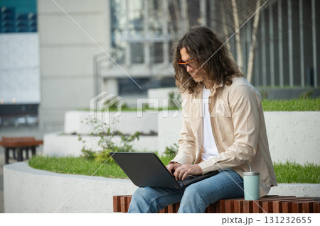 Handsome man, attractive freelancer , brunette sits on bench in urban park, using laptop for remote work. Wearing casual clothes. Outdoors. freelance Handsome man, attractive freelancer , brunette sits on bench in urban park, using laptop for remote work. Wearing casual clothes. Outdoors. freelance 131232655