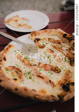 Round pizza with bacon and chives is being sliced with pizza cutter on white plate on wooden table, with slice already served on another plate. Crispy bacon and chives pizza slicing on white plate 131232967