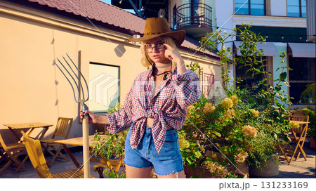 Stylish woman in cowboy hat holding pitchfork with confident attitude in urban garden 131233169