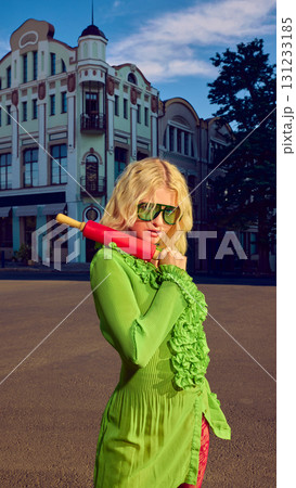 Blonde woman in green dress holding red rolling pin with bold confidence on street Blonde woman in green dress holding red rolling pin with bold confidence on street 131233185