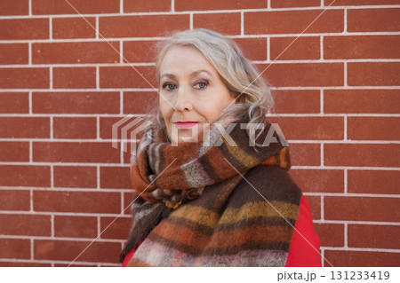Gray-haired woman of sixty years old in a red coat and a terracotta scarf near a brick wall 131233419