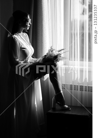 Bride in a white dress, holding a bouquet, sits by a window, lost in thought. 131235337
