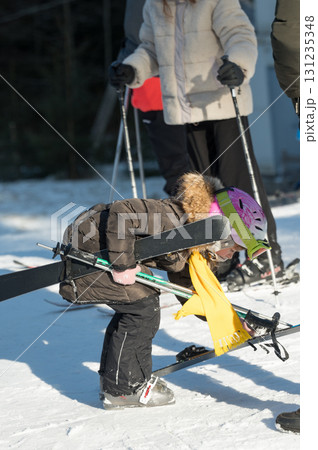 Little skier struggles to lift equipment on snowy mountain slope while surrounded by supportive adults during a sunny winter day Little skier struggles to lift equipment on snowy mountain slope while surrounded by supportive adults during a sunny winter day 131235348