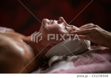 Side view close up of a mature woman lying on a treatment bed with a headband while a professional therapist massages her face. 131236509