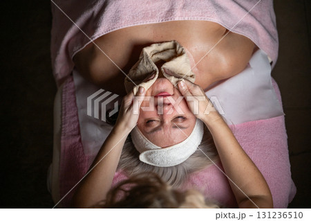 Close up of a mature woman lying on a treatment bed with a towel and headband while a professional therapist applies foam during a facial cleansing procedure. Skincare and wellness concept Close up of a mature woman lying on a treatment bed with a towel and headband while a professional therapist applies foam during a facial cleansing procedure. Skincare and wellness concept 131236510