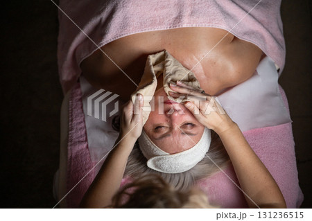 Close up of a mature woman lying on a treatment bed with a towel and headband while a professional therapist applies foam during a facial cleansing procedure. Skincare and wellness concept 131236515