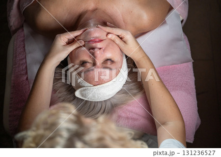 Close up of a mature woman receiving a relaxing facial massage from a professional female therapist in a modern spa. The client lies on a treatment bed with a towel and headband, enjoying skincare 131236527