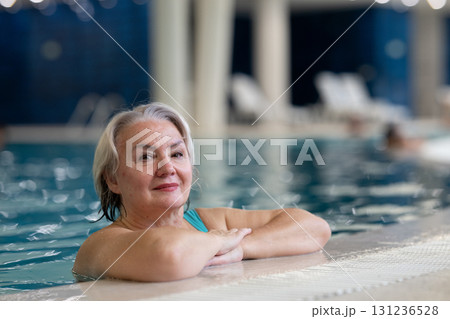 Smiling senior woman enjoying relaxation in an indoor swimming pool, leaning on the poolside. Concept of active lifestyle, wellness, health, hydrotherapy, and leisure for older adults. 131236528