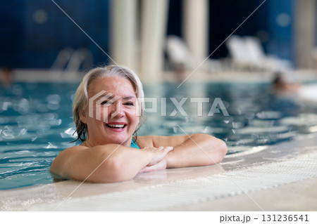 Smiling senior woman enjoying relaxation in an indoor swimming pool, leaning on the poolside. Concept of active lifestyle, wellness, health, hydrotherapy, and leisure for older adults. Smiling senior woman enjoying relaxation in an indoor swimming pool, leaning on the poolside. Concept of active lifestyle, wellness, health, hydrotherapy, and leisure for older adults. 131236541